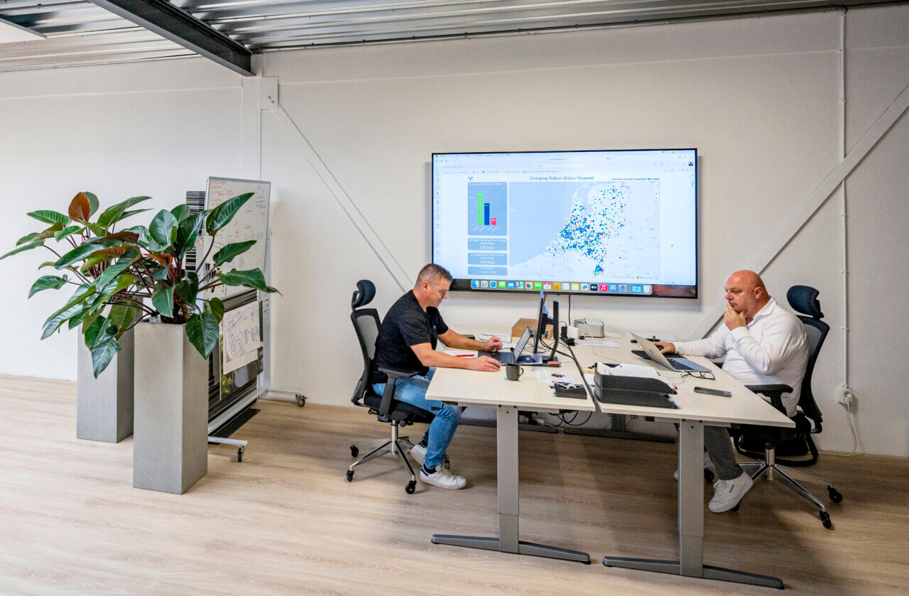 Collega's Tommy en René zijn aan het werk op kantoor. Naast hun hangt een groot tv-scherm waarop het NOC te zien is.