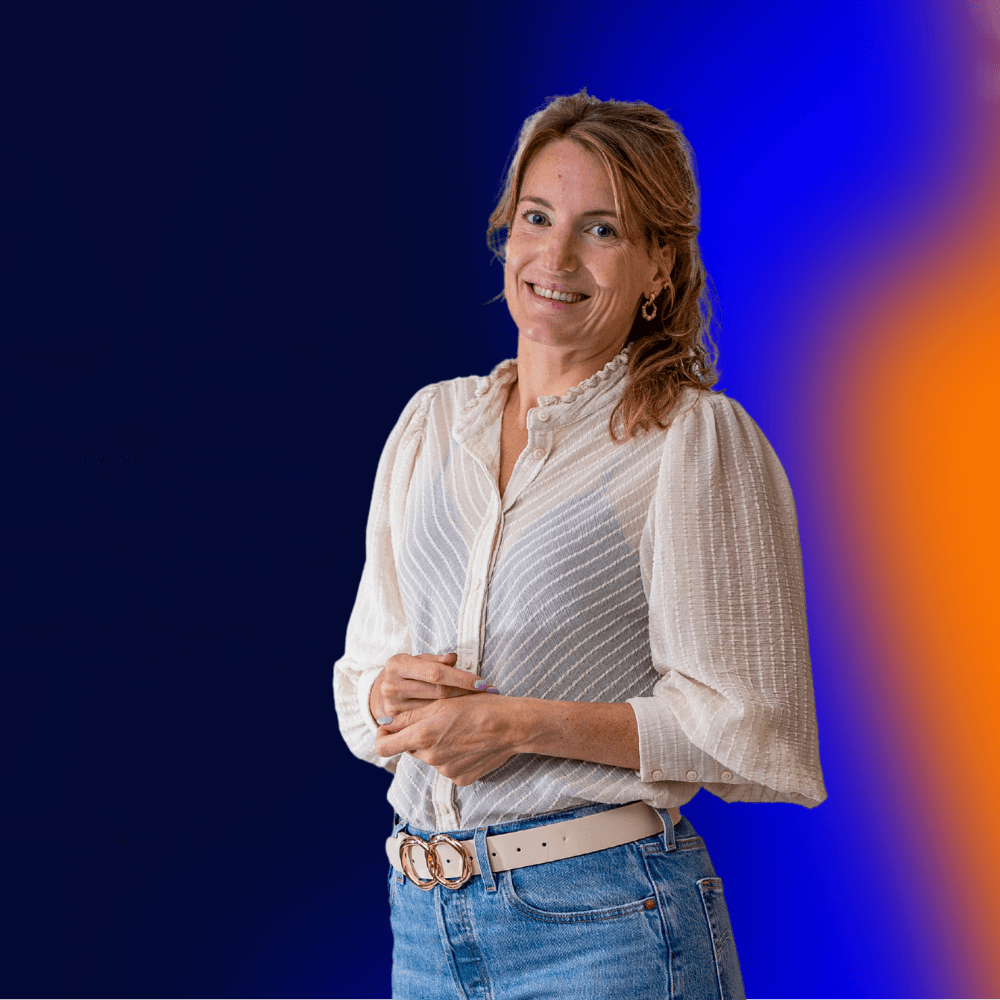 Vrouw staat met handen in elkaar gevouwen voor een blauwe achtergrond. Ze lacht vriendelijk.